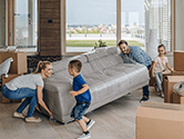 A man and woman lift a couch to move while their children run around the room