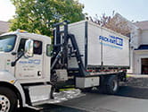 View of a 1-800-PACK-RAT driver backing in a mini-mover truck with a portable storage container on the back of it into the driveway of a customer as the customers watch on a bright sunny day