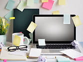 A cluttered work desk where a laptop with sticky notes on it sits