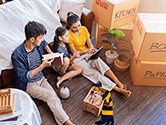 A family sits on the floor of their new home looking at a tablet surrounded by marked packed boxes