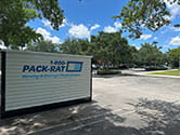 A side view of a 1-800-PACK-RAT portable storage container sitting in the parking lot of a business on a bright sunny day