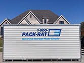 Side view of a 16-foot 1-800-PACK-RAT portable storage container that is being delivered to the street-front of a suburban home on a bright sunny day