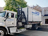 A 1-800-PACK-RAT mini-mover truck drops off a portable storage container in the driveway of a suburban home with the family watching