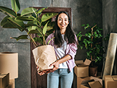 A woman smiles as she holds her houseplant in her hands surrounded by packed and open boxes inside her home