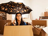 A woman sits in a box with an umbrella as she unpacks her belongings